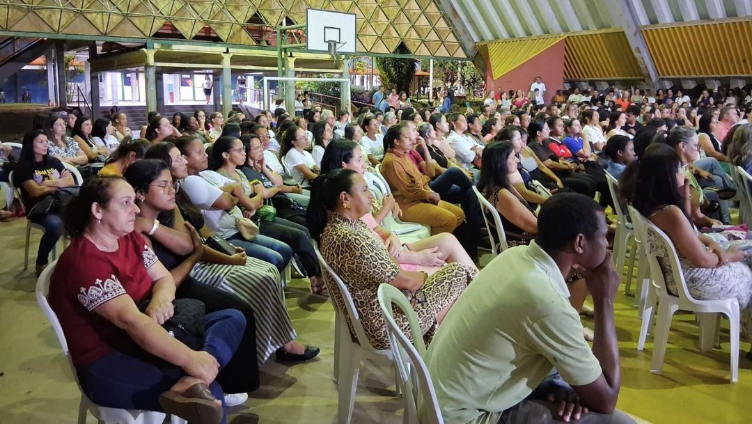 Vista de perfil de um grande público sentado em cadeiras brancas em uma quadra poliesportiva. A maioria das pessoas são mulheres, atentas ao evento. Ao fundo, vê-se a estrutura metálica do teto da quadra e uma tabela de basquete.
