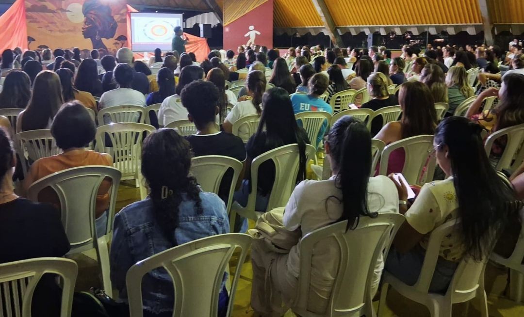 Vista de costas do público sentado em direção ao palco. No palco, há um telão exibindo um slide e uma pintura artística ao fundo representando uma mulher negra com turbante em uma paisagem de savana ao pôr do sol. Um palestrante fala ao microfone no canto esquerdo.