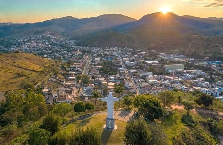 Vista aérea de Guaçuí com monumento do Cristo em primeiro plano, tendo ao fundo montanhas onde se reflete um por de sol.