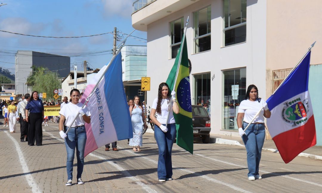 Três jovens caminham em um desfile cívico segurando as bandeiras do estado do Espírito Santo, do Brasil e do município de Dores do Rio Preto. Elas vestem calça jeans e camiseta branca. Ao fundo, o público acompanha o trajeto em uma rua pavimentada.