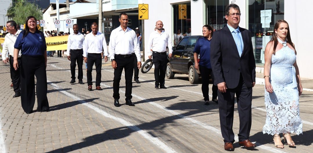 Autoridades municipais, incluindo o prefeito Thiaguinho, a primeira-dama Alexandra e vereadores, caminham em formação durante o desfile cívico em Dores do Rio Preto. Estão vestidos com trajes formais e casuais finos sobre uma rua de blocos sextavados, sob céu claro.