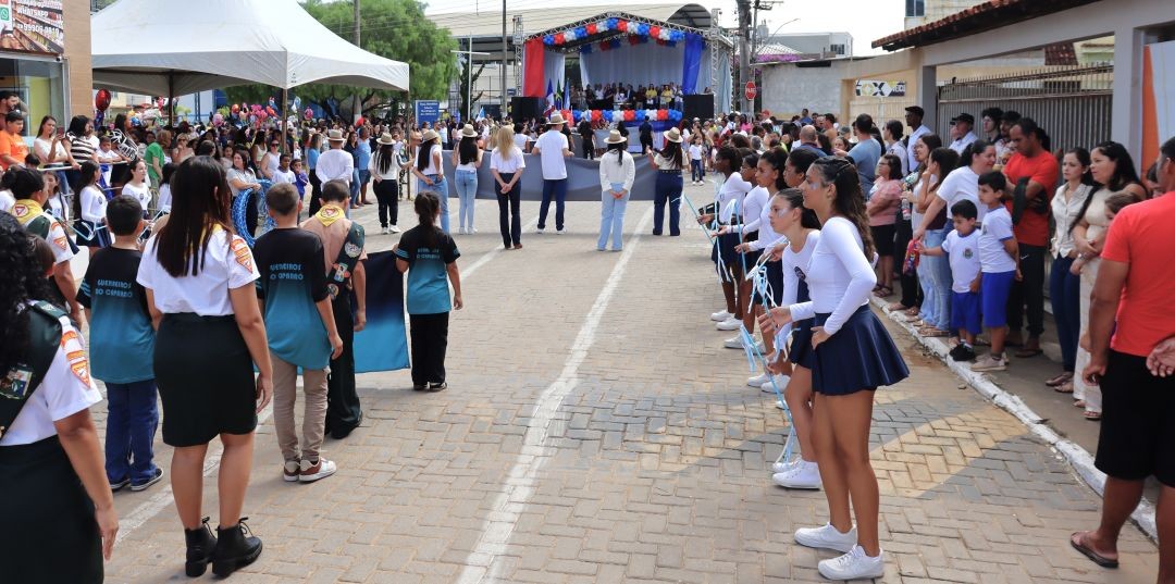 Visão geral da Rua Adair Furtado de Souza durante o desfile. Grupos organizados ocupam o centro da via pavimentada, incluindo jovens com uniformes e as fitas rítmicas de balizas. Ao fundo, vê-se um palco montado e decorado com cores cívicas. O público acompanha a movimentação nas calçadas laterais.