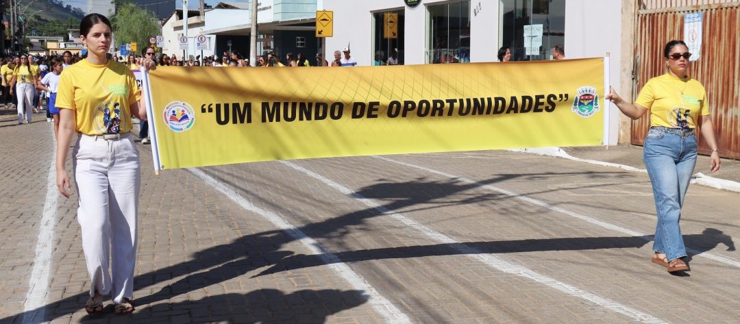Duas mulheres desfilam em primeiro plano, segurando um grande banner amarelo. O banner exibe a frase em destaque: “UM MUNDO DE OPORTUNIDADES”, ladeada por logotipos institucionais. Ambas vestem camisetas amarelas do evento; a mulher à esquerda usa calça branca e a da direita, calça jeans. Elas caminham sobre uma rua pavimentada, seguidas por outros participantes ao fundo.