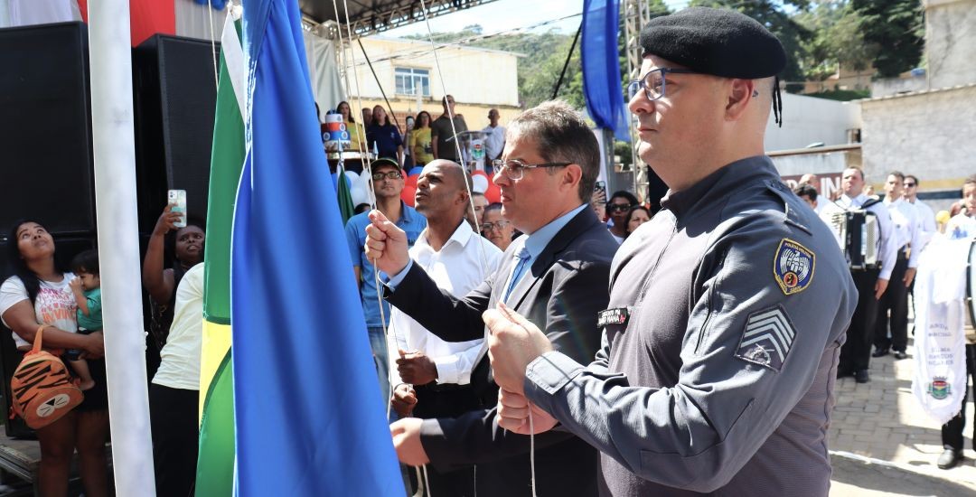 Detalhe da solenidade de hasteamento das bandeiras durante o aniversário de Dores do Rio Preto. O prefeito Thiaguinho, de terno, puxa o cordão da bandeira azul, acompanhado por um homem de camisa branca e um sargento da Polícia Militar fardado e com boina preta. O sargento segura a bandeira azul. No fundo, vê-se um palco decorado, pessoas na audiência e músicos da banda marcial com instrumentos. Uma mulher segura um bebê no colo à esquerda.