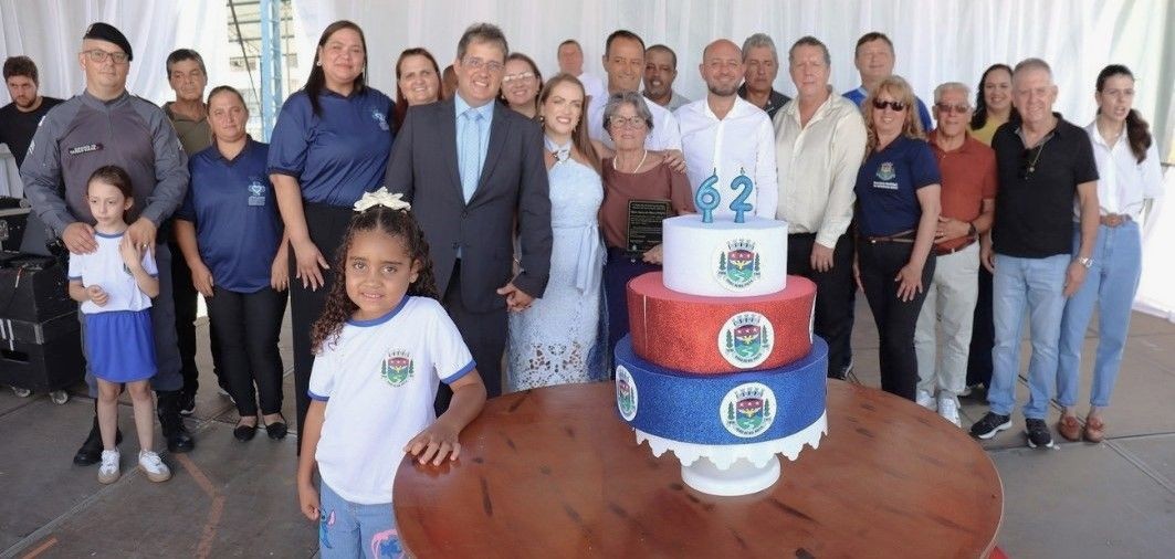 Um grupo de pessoas está reunido para uma foto oficial atrás de um bolo de aniversário de três andares. O bolo é decorado nas cores azul, vermelha e branca, com o brasão do município e o número 
