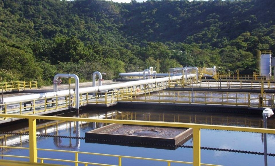 Estação de tratamento de esgoto com tanques, tubulações e passarelas metálicas, cercada por área verde e relevo montanhoso ao fundo.