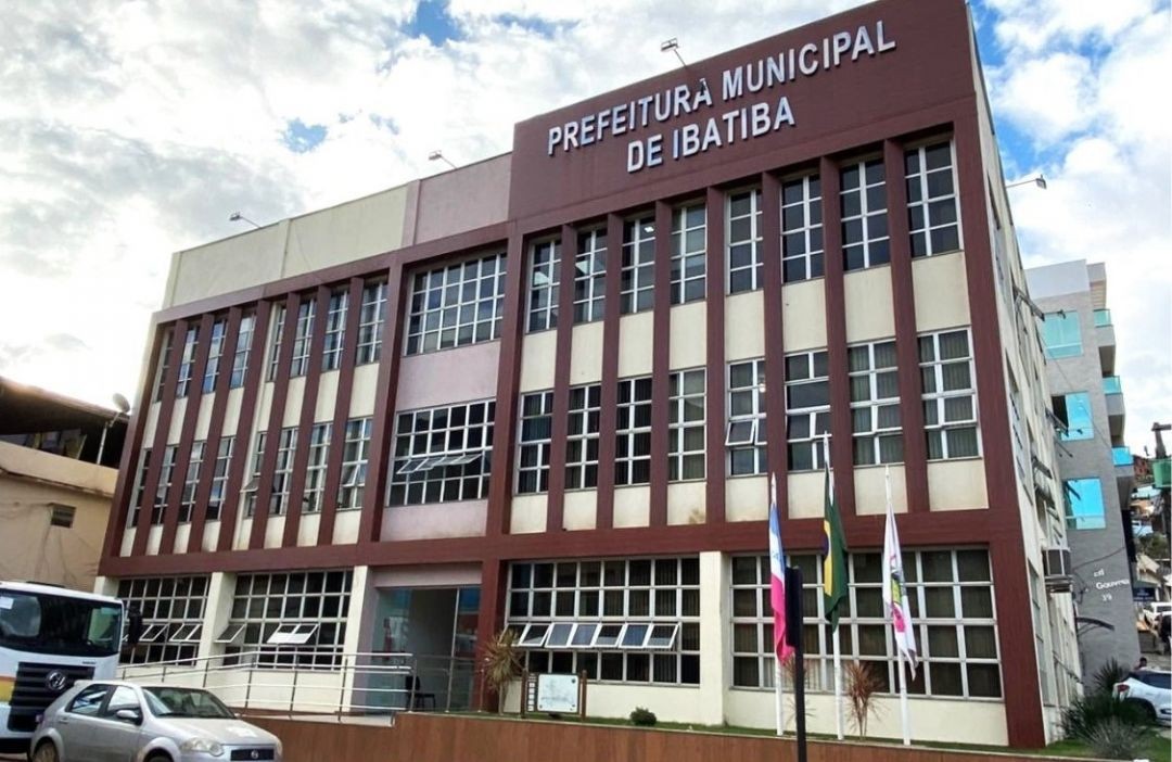 Fachada frontal do prédio da Prefeitura Municipal de Ibatiba. A edificação possui três pavimentos, com janelas de vidro e detalhes em colunas de cor marrom. No topo, destaca-se o letreiro em letras brancas com o nome do órgão. Em frente à entrada, há três mastros com as bandeiras do Espírito Santo, do Brasil e do município, além de veículos estacionados na via pública.