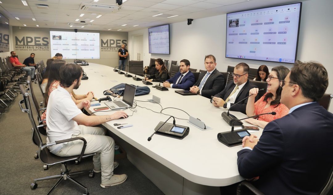 1.	Sala de reuniões do MPES com integrantes em volta de uma mesa longa durante encontro institucional, com telas digitais ao fundo.