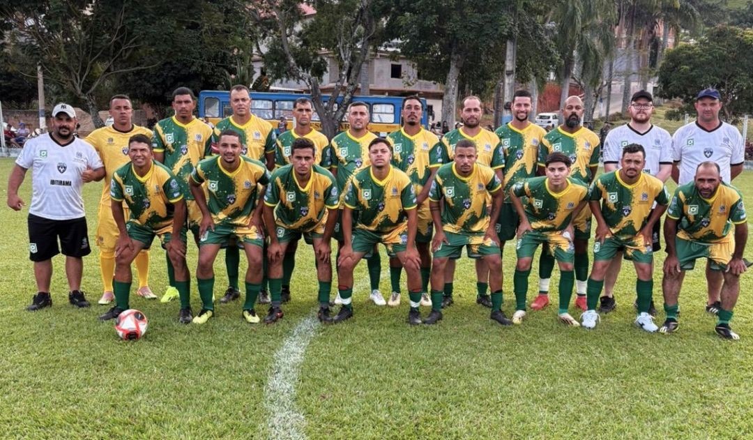 Fotografia colorida da equipe masculina de Ibitirama posada em um campo de futebol gramado. Os jogadores vestem uniforme verde e amarelo com detalhes em branco. Ao fundo, árvores e um ônibus azul de transporte escolar.