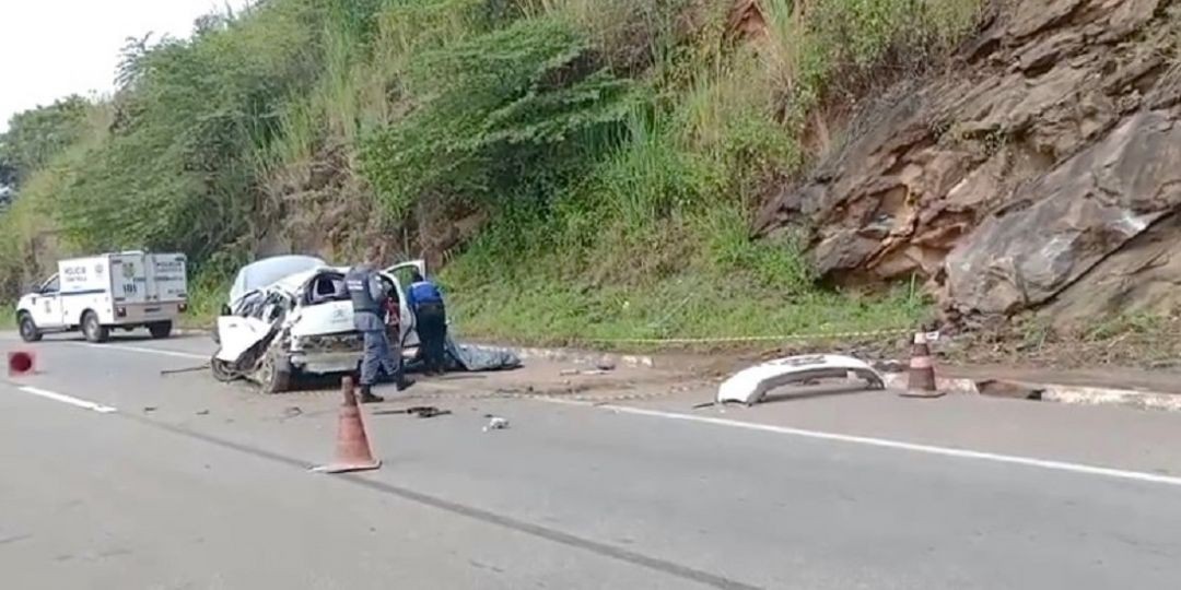 Imagem traseira de carro branco destruído após colisão, na Rodovia ES-482, contra uma ribanceira – em destaque no lado direito da foto, onde também se vê o para-choque do veículo, cones de sinalização na pista e equipes atuando no local do acidente, tento ao fundo uma viatura da Polícia Científica.