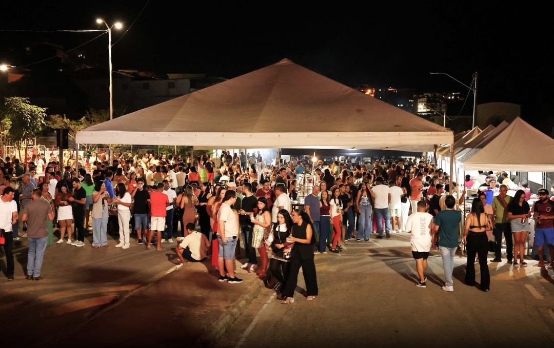 Imagem noturna de uma praça movimentada com uma grande tenda branca centralizada. Diversas pessoas circulam pelo espaço iluminado por postes de luz.