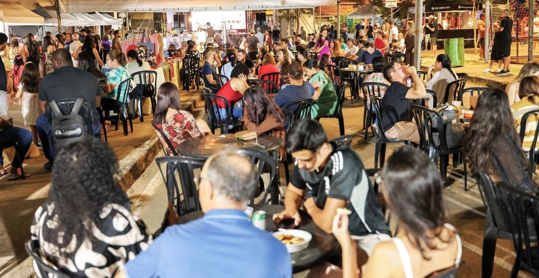 Visão de pessoas sentadas em mesas e cadeiras de plástico pretas sob uma estrutura coberta. Ao fundo, é possível ver o palco e outras barracas de alimentação.