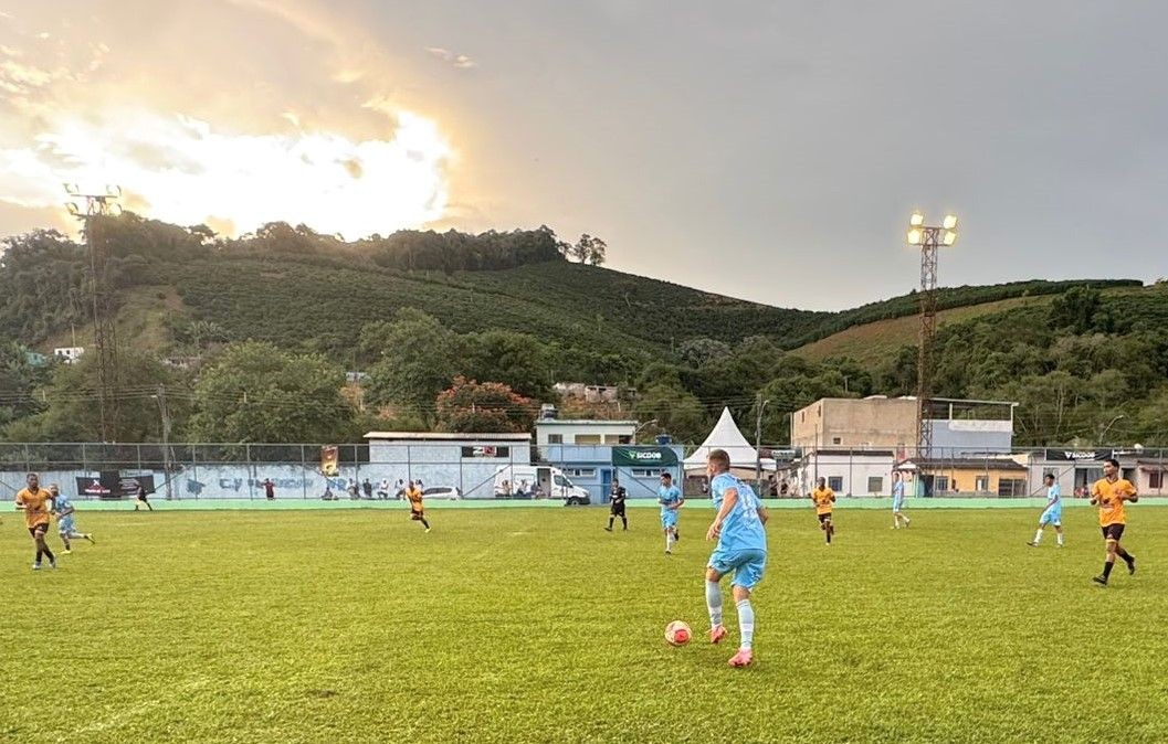 Jogadores disputam partida de futebol em campo gramado no Estádio Municipal de Dores do Rio Preto, com equipes uniformizadas em ação durante o campeonato regional ao entardecer.
