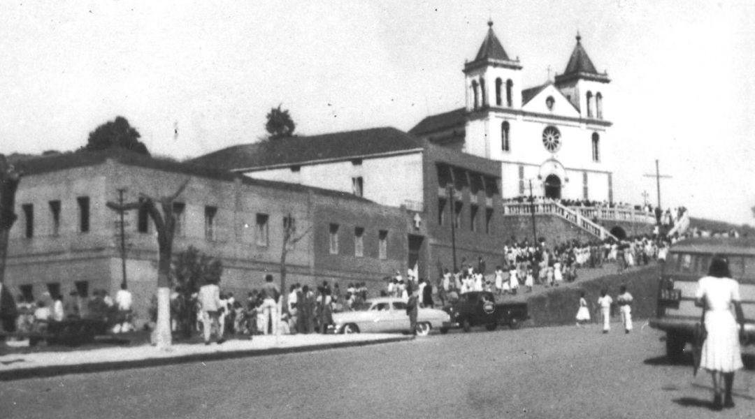 Fotografia histórica da fachada da Igreja Matriz de Alegre com uma grande quantidade de fiéis na escadaria e na praça. Ao fundo, carros antigos da década de 50 estacionados na rua lateral.