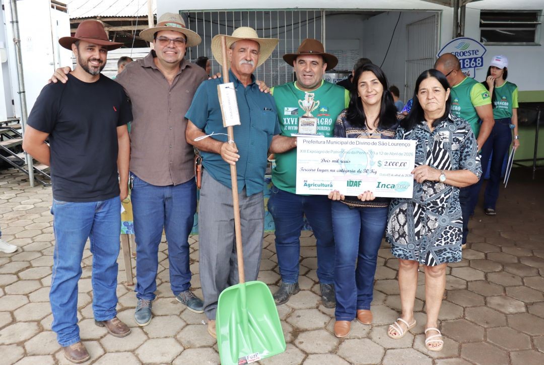 Três homens e duas mulheres posam para a foto em uma área externa pavimentada. Um senhor ao centro segura uma pá de plástico verde e um troféu, enquanto as mulheres seguram um cheque simbólico de 2 mil reais. O grupo está sorridente e veste trajes casuais e chapéus.