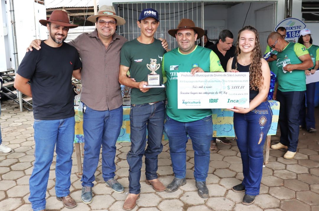 Três homens e uma jovem posam juntos. O homem ao centro veste uma camisa verde com a logomarca do evento e segura um troféu dourado. A jovem à direita e o homem ao centro seguram um cheque simbólico no valor de 3 mil reais.