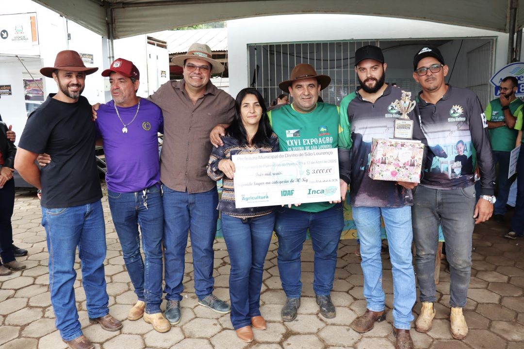 Grupo de seis pessoas posa para registro da premiação. Dois homens à direita vestem camisas de equipe na cor cinza com detalhes verdes. Ao centro, uma mulher e um homem seguram um cheque simbólico de 3 mil reais. Um dos integrantes segura um troféu e uma caixa de presente decorada.