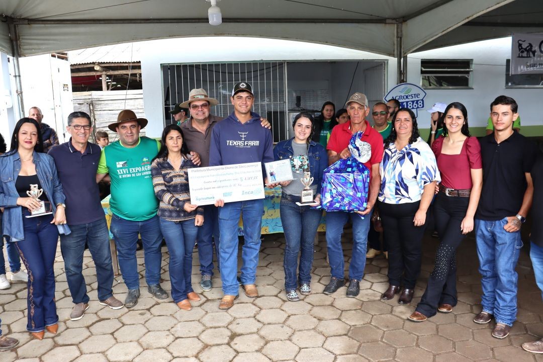 Um grupo maior, composto por cerca de dez pessoas, posa sob a tenda do evento. No centro, um jovem com moletom azul segura um cheque simbólico de 4.500 reais. Ao lado, uma jovem segura um troféu e um homem segura um grande presente embrulhado em papel azul brilhante.