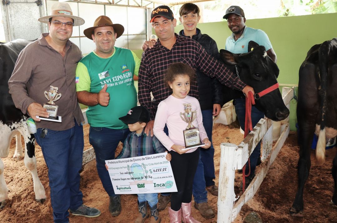 Três homens e duas crianças posam ao lado de uma vaca preta em um curral. Uma menina segura um troféu e um cheque simbólico no valor de 6 mil reais da Prefeitura de Divino de São Lourenço.