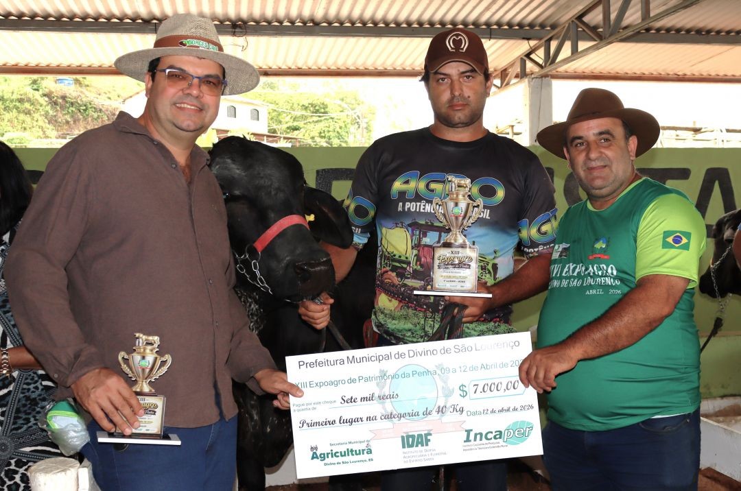 Três homens posam segurando um cheque simbólico de 7 mil reais e um troféu dourado. Ao fundo, aparece a cabeça de uma vaca de pelagem escura. Eles vestem roupas casuais e chapéus de aba larga.
