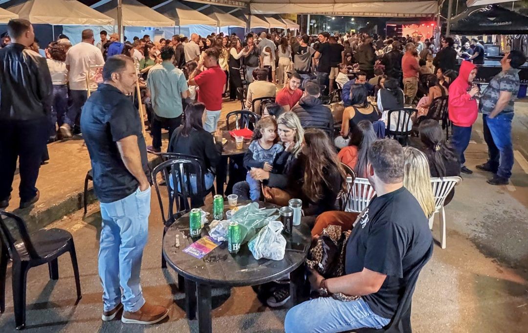 Público sentado em mesas de plástico pretas em uma área aberta da feira sob tendas. No primeiro plano, uma mesa com latas de cerveja e embalagens de comida, enquanto ao fundo várias pessoas conversam e circulam entre as barracas.