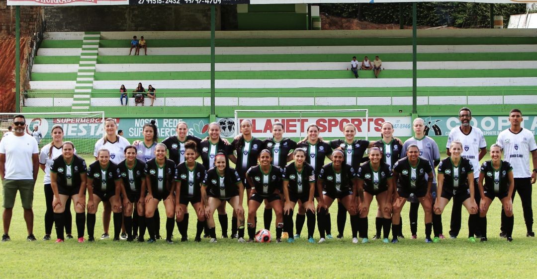 Equipe feminina de futebol de Afonso Cláudio posa alinhada no campo antes da partida, com atletas uniformizadas em preto e verde, acompanhadas por comissão técnica, em arquibancada ao fundo.