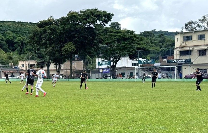 Jogadores disputam partida de futebol em campo gramado no Estádio Municipal de Dores do Rio Preto, com equipes uniformizadas em ação durante o campeonato regional tendo ao fundo um alambrado com faixas do “Sicoob”, onde estão algumas pessoas assistindo e casas da cidade.