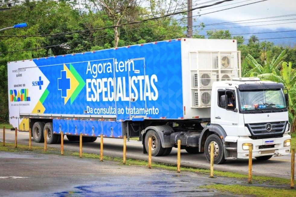Fotografia em ângulo lateral de um caminhão branco da marca Mercedes-Benz puxando um baú refrigerado de grande porte. A lateral do baú é azul com detalhes em formas geométricas e traz a identidade visual do Governo Federal e do SUS. Destaca-se o texto em letras brancas grandes: 