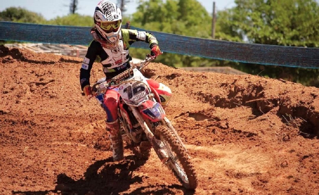 Piloto de motocross, com o número 16, faz curva em pista de terra, tendo ao fundo uma faixa azul, vegetação verde e dia ensolarado.