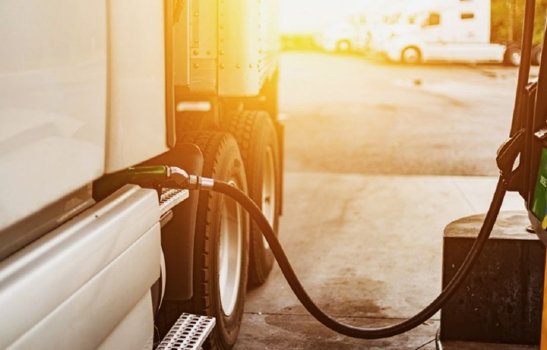 Fotografia em close lateral de um caminhão branco sendo abastecido em um posto de combustível. A mangueira de diesel está conectada ao tanque do veículo. Ao fundo, o pátio do posto está iluminado por uma luz solar intensa, com outros caminhões estacionados de forma desfocada.