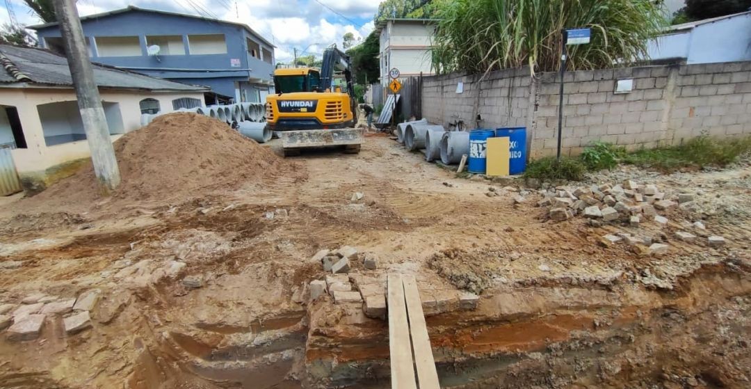 Rua em obra com escavadeira, tubos de concreto para drenagem e solo preparado para futura pavimentação.