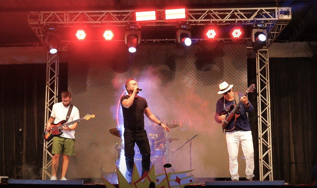 Banda de pop-rock se apresenta em palco iluminado com refletores coloridos e telão de LED ao fundo. Três músicos aparecem: um baixista, um vocalista ao centro e um guitarrista de chapéu à direita.