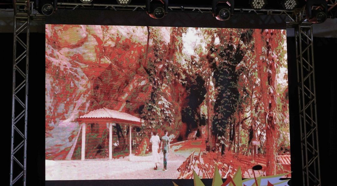 Detalhe de um grande telão de LED em uma estrutura de metal, exibindo a cena de um filme onde duas pessoas caminham em um ambiente arborizado próximo a um quiosque.