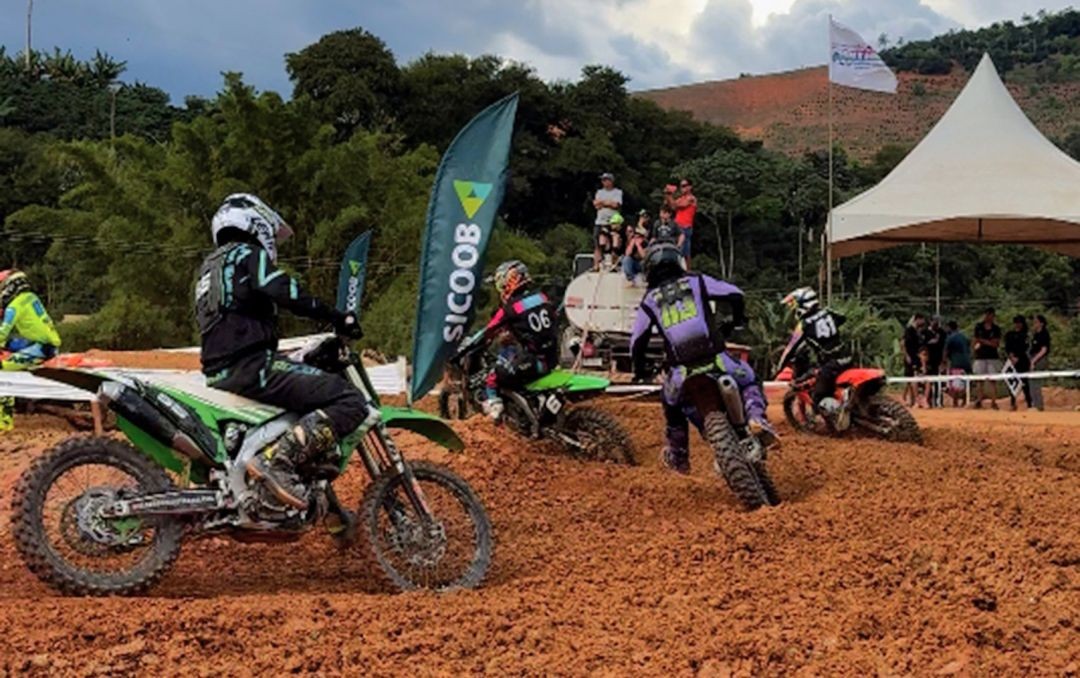 Pilotos de motocross em uma curva acentuada de terra batida durante competição em Iúna. Ao fundo, bandeiras de patrocinadores e público assistindo sob tendas.