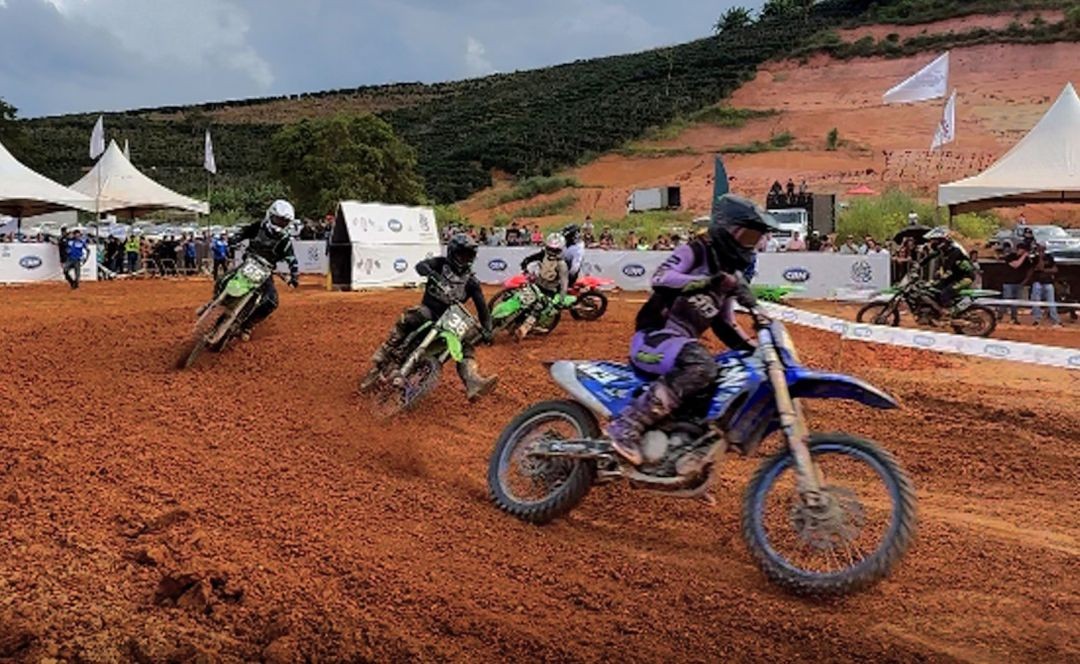 Competidores acelerando suas motocicletas em uma reta de terra cor de laranja. É possível ver a vegetação de cafezais ao fundo nas encostas.