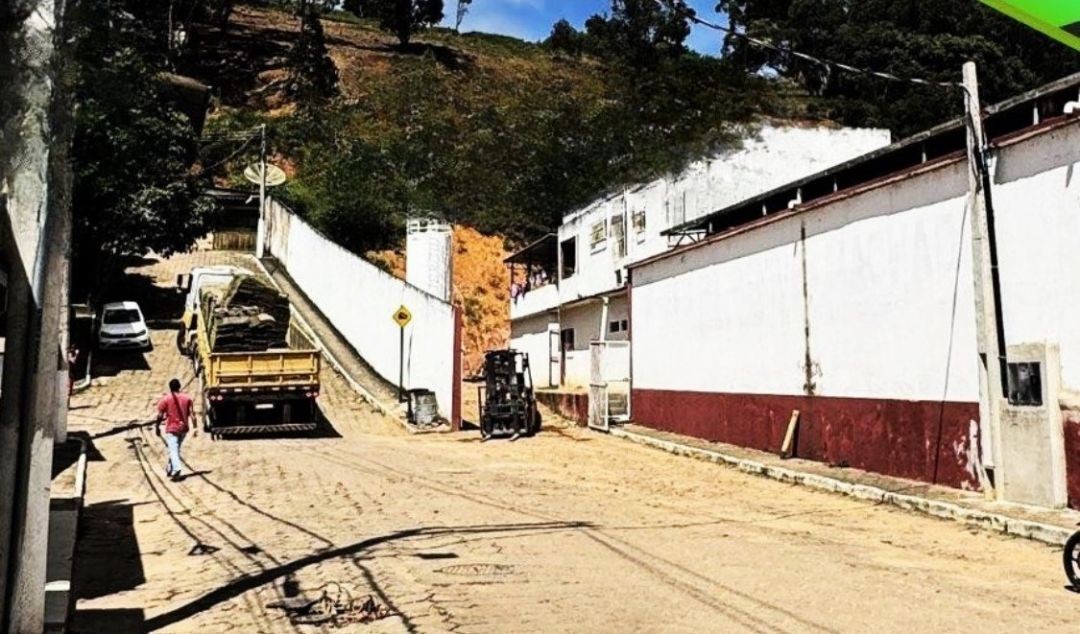 Rua em aclive em Irupi com caminhão de obras estacionado, solo parcialmente escavado e trecho com interdição durante serviços de infraestrutura próximos a um prédio e área urbana.