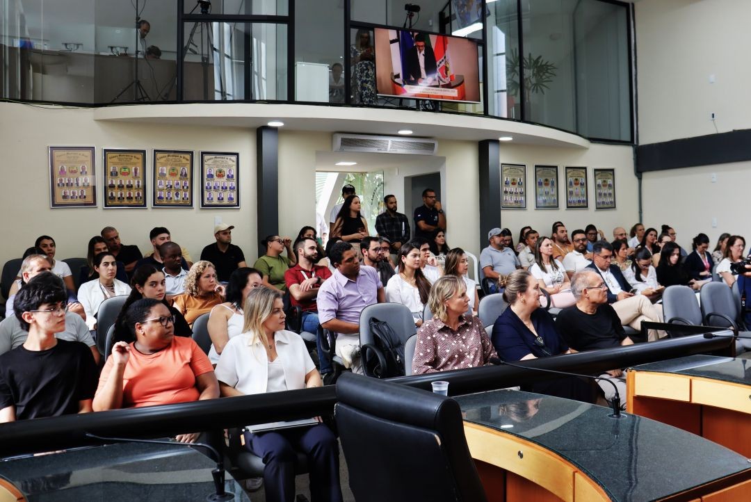 Foto apresenta, em primeiro plano, o público composto por servidores e gestores assistindo à cerimônia de abertura do Enfoc 2026, tendo ao fundo parede com quadros e pessoas em pé na entrada do prédio. Acima, uma galeria de vidro e uma tela de vídeo que apresenta o orador.