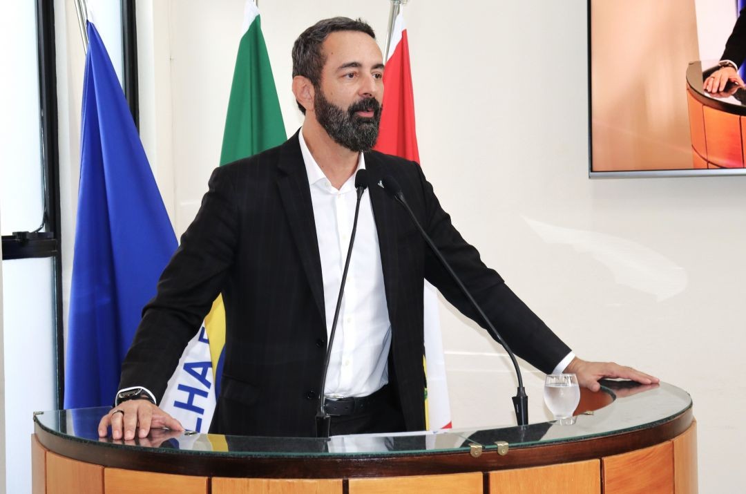 Fotografia em plano médio do conselheiro Rodrigo Chamoun, homem de barba e terno preto, falando ao púlpito de madeira e vidro. Ao fundo, as bandeiras do Brasil, do Espírito Santo e do município.