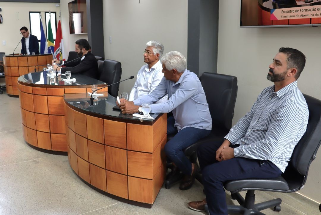 Autoridades sentadas à mesa diretora da Câmara Municipal acompanham o evento. Entre eles, homens de camisas sociais clara e escura, em um ambiente formal e iluminado.
