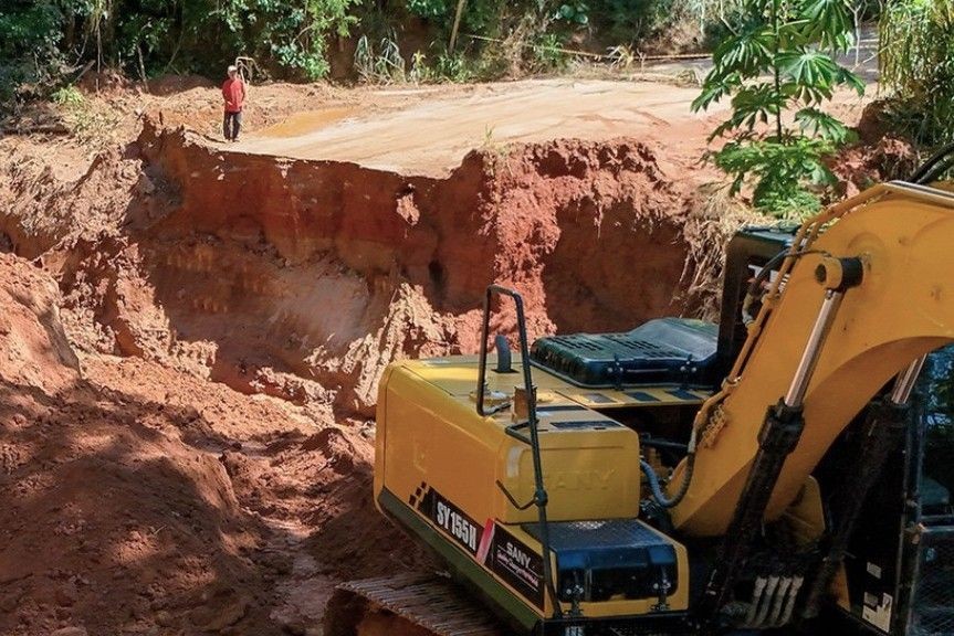 Escavadeira trabalha na recuperação de estrada rural com grande cratera aberta após rompimento de bueiro, enquanto um homem observa o local interditado.