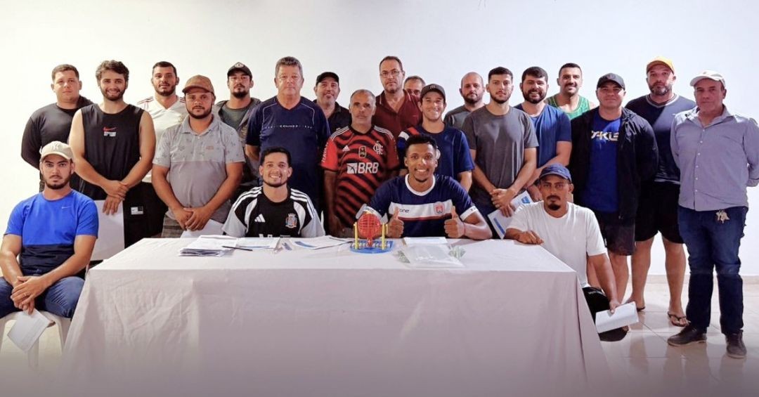 Grupo de aproximadamente 20 homens posados para foto atrás de uma mesa coberta com toalha branca. Ao centro da mesa, há um pequeno globo de sorteio e documentos. Eles representam as equipes do Torneio do Trabalhador de Iúna.