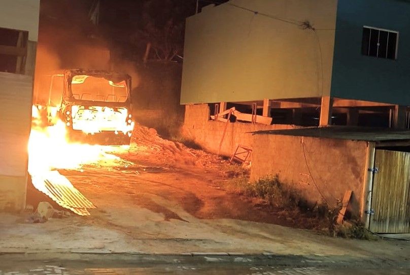 Fotografia noturna mostra um ônibus de grande porte sendo consumido por chamas intensas em um terreno baldio ao lado de uma edificação em construção. O fogo ilumina o chão e as paredes próximas.
