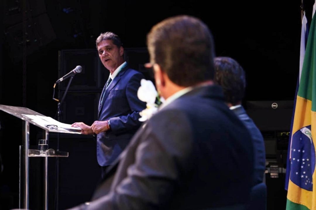 A fotografia mostra, no lado esquerdo, o governador do Espírito Santo, Ricardo Ferraço, vestindo terno azul e camisa social, em pé e falando ao microfone em um púlpito transparente. Ele segura folhas de papel e olha para a direita. Em primeiro plano, no lado direito e de costas para a câmera, estão a silhueta e as costas de outro homem, sentado e com terno azul-marinho. Ao fundo, à direita, vê-se a bandeira do Brasil.