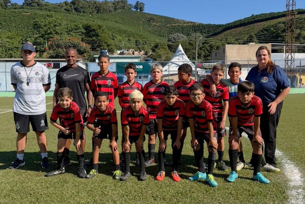 Fotografia colorida de um time de futebol juvenil em um campo de grama natural sob luz solar. Os atletas mirins vestem uniformes listrados em preto e vermelho (horizontal). À esquerda e à direita, estão três adultos (comissão técnica): um homem de camisa polo branca, um homem de camisa preta e uma mulher de camisa polo azul marinho. Ao fundo, uma encosta com vegetação densa e cafezais sob um céu azul.