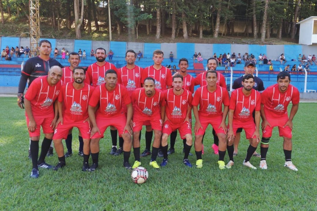 Fotografia de um time de futebol amador posicionado para foto oficial no gramado. Os jogadores vestem uniforme vermelho com listras verticais brancas finas e calções vermelhos. Ao fundo, arquibancada azul com torcedores e área arborizada.