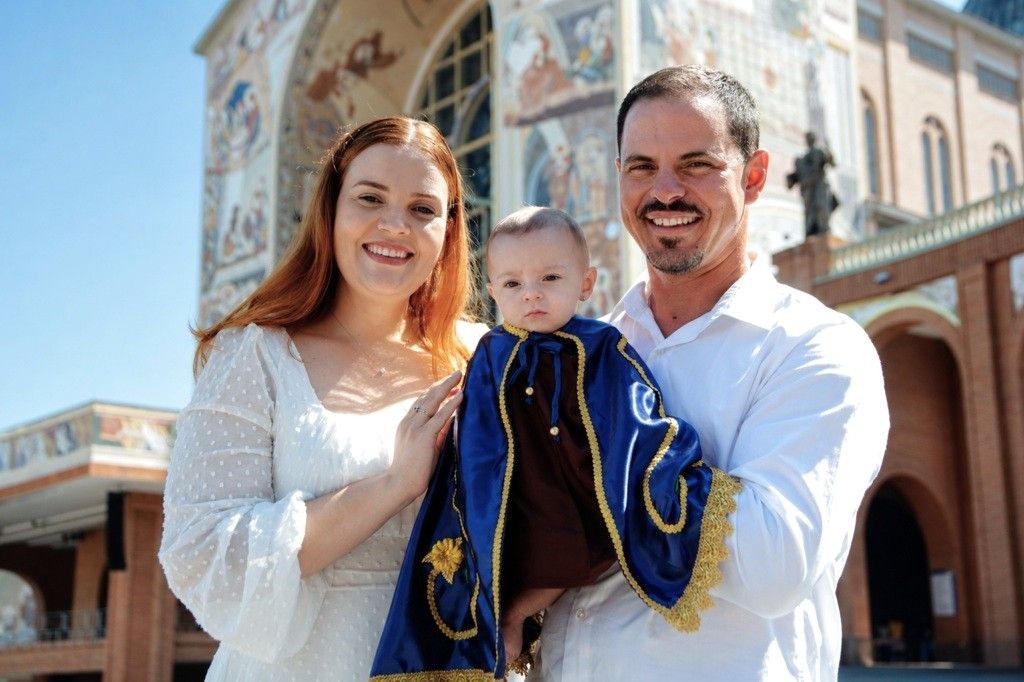 Retrato em plano médio da mãe Bruna, o pai, Jhonatan e a bebê Eva em frente à fachada do Santuário Nacional de Aparecida. O pai segura a bebê que veste uma capa azul com bordados dourados, semelhante ao manto de Nossa Senhora Aparecida. Todos sorriem sob a luz do sol.