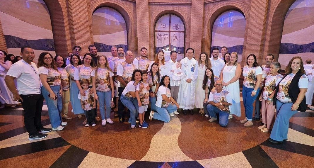 Foto em grupo com dezenas de familiares e amigos no interior de uma capela circular com paredes de tijolos aparentes. Ao centro, um padre posa ao lado dos pais e da bebê. Muitos convidados vestem camisetas personalizadas com a foto da pequena Eva e frases religiosas.