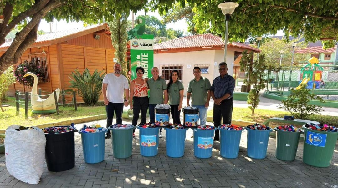 Servidores da Semmam e voluntários posam atrás de baldes contendo tampinhas plásticas, antes d serem encaminhadas para reciclagem, tendo ao fundo um jardim e duas casas de alvenaria e de madeira, sede da Secretaria de Meio Ambiente.