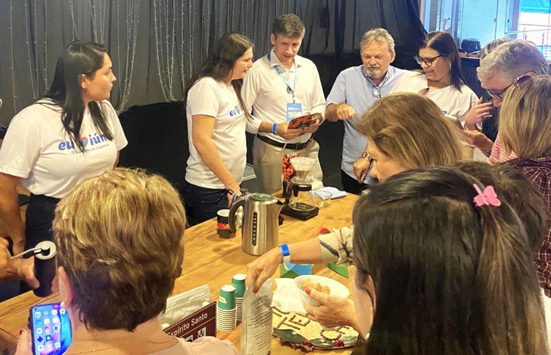 Vista superior da movimentação no estande de Iúna. Produtores e visitantes interagem ao redor da mesa de degustação. É possível ver o momento do preparo do café com métodos de filtragem manual e pessoas observando materiais informativos.