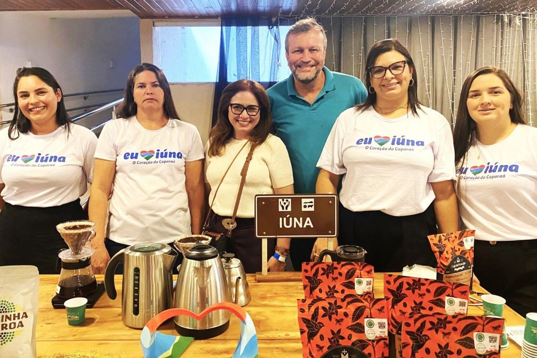 Cinco mulheres e um homem posam sorridentes em frente ao balcão de exposição. No centro, uma placa marrom identifica o município de 